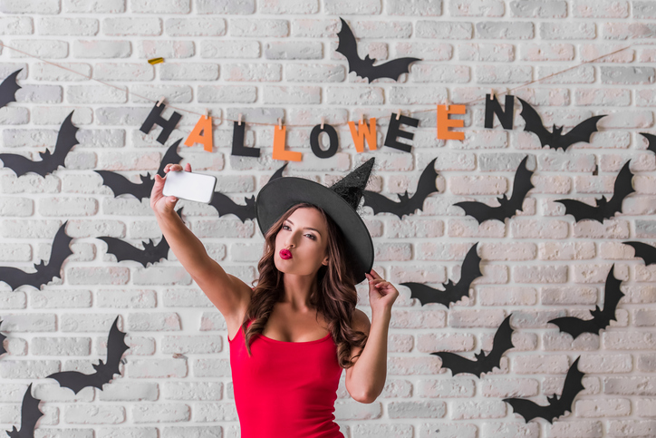 Beautiful sexy girl in red dress and witch hat is doing selfie using a smart phone, on background decorated for Halloween