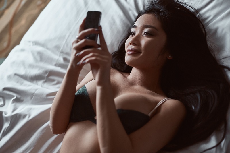 Portrait of a beautiful sensual Asian woman in underwear lying on the bed with the phone in his hand. Interior shot