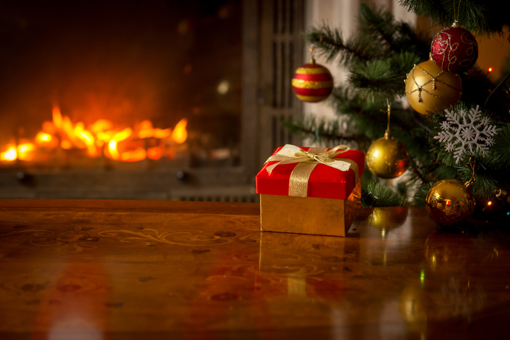 Closeup image of red gift box on wooden table in front of burning fireplace and Christmas tree